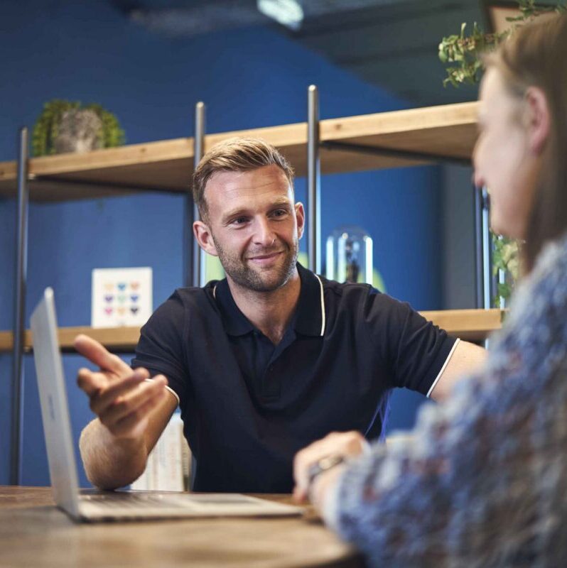 Man speaking to woman at a table with a laptop
